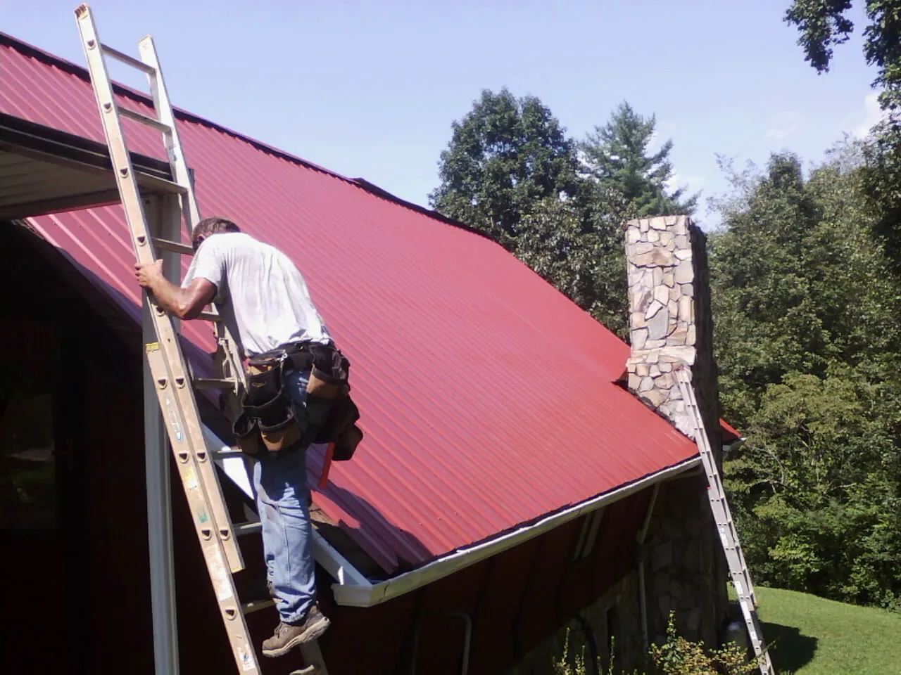 Roofer on a ladder beside a red metal roof and stone chimney — Soffit & Fascia Repair by Roofing Brothers in Tumtum